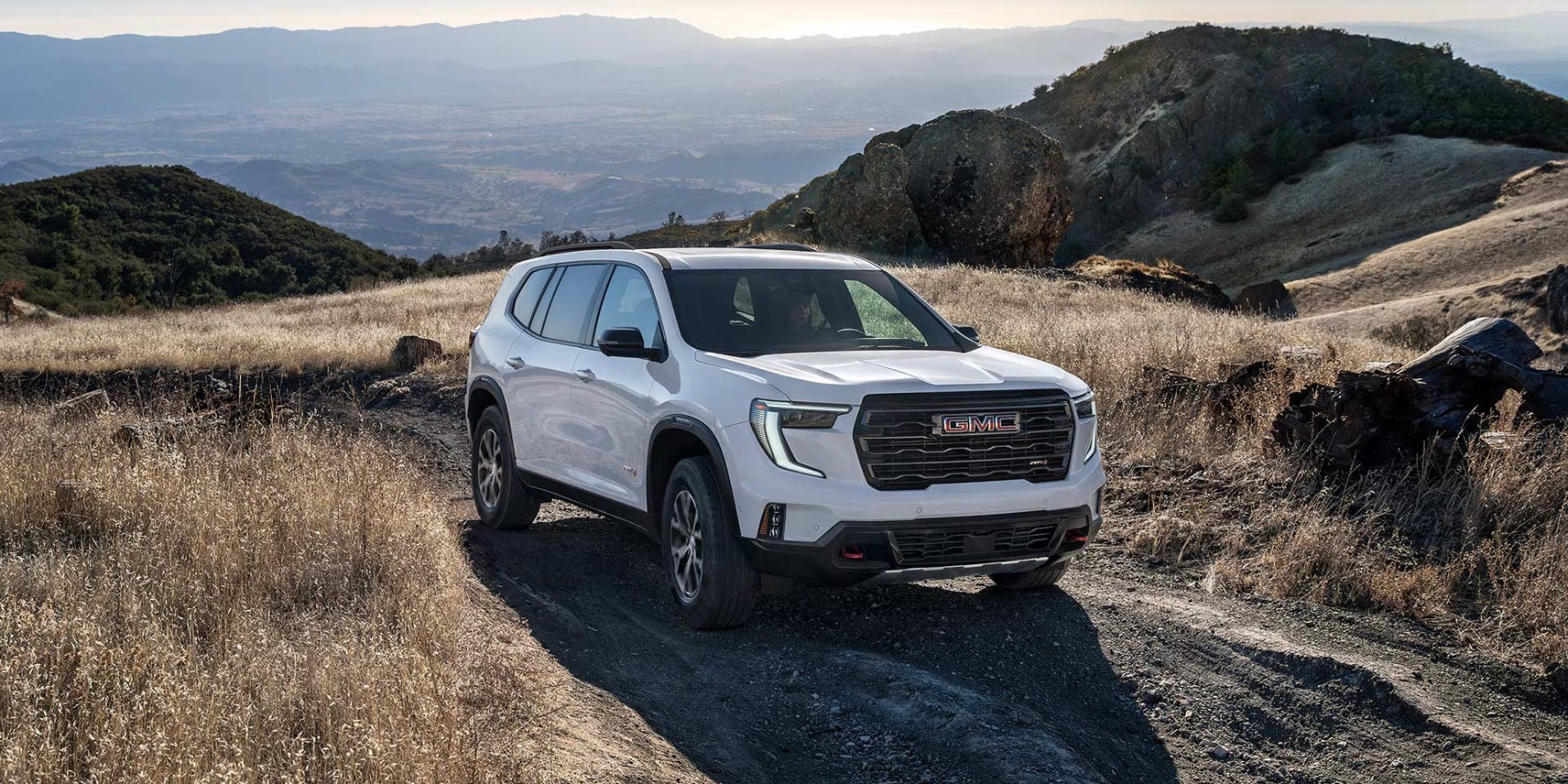 A white GMC Acadia driving on a dirt road in a hilly field.