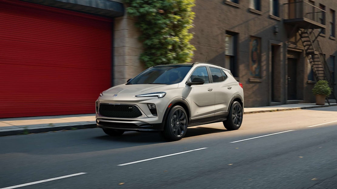 A light-colored Buick Encore GX driving down the street in a town.