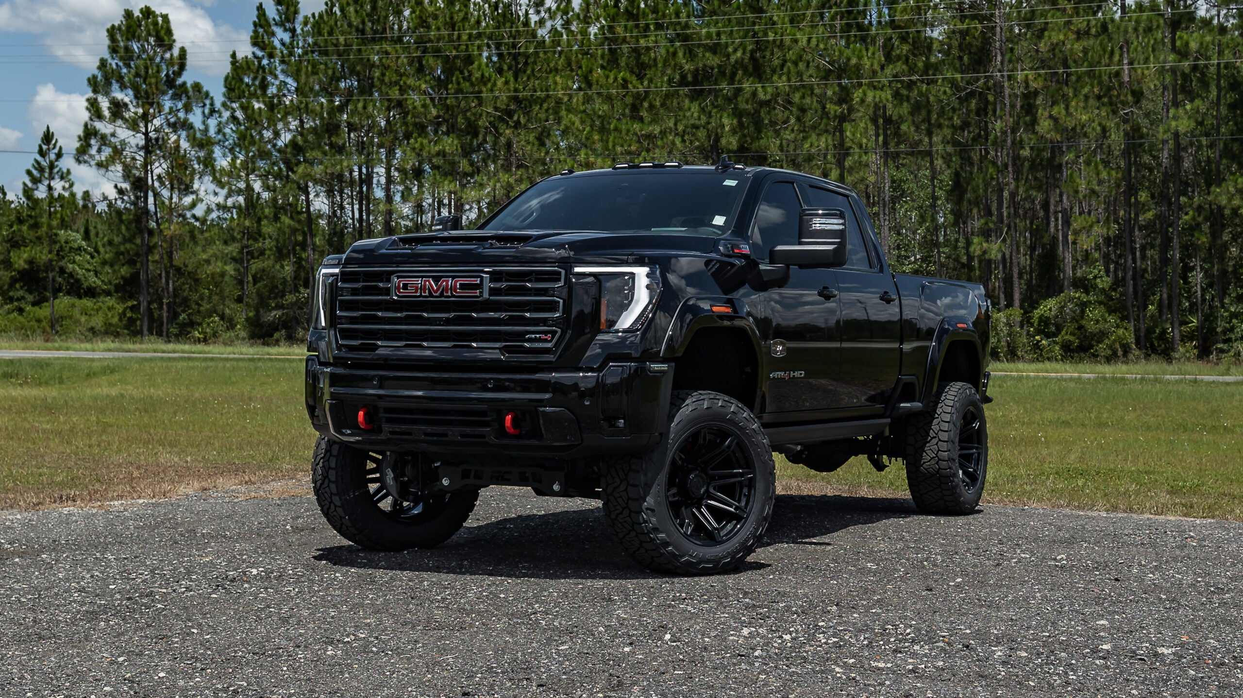 Black lifted GMC Sierra HD pickup truck with large off-road wheels and red tow hooks, parked on pavement with pine trees in the background.