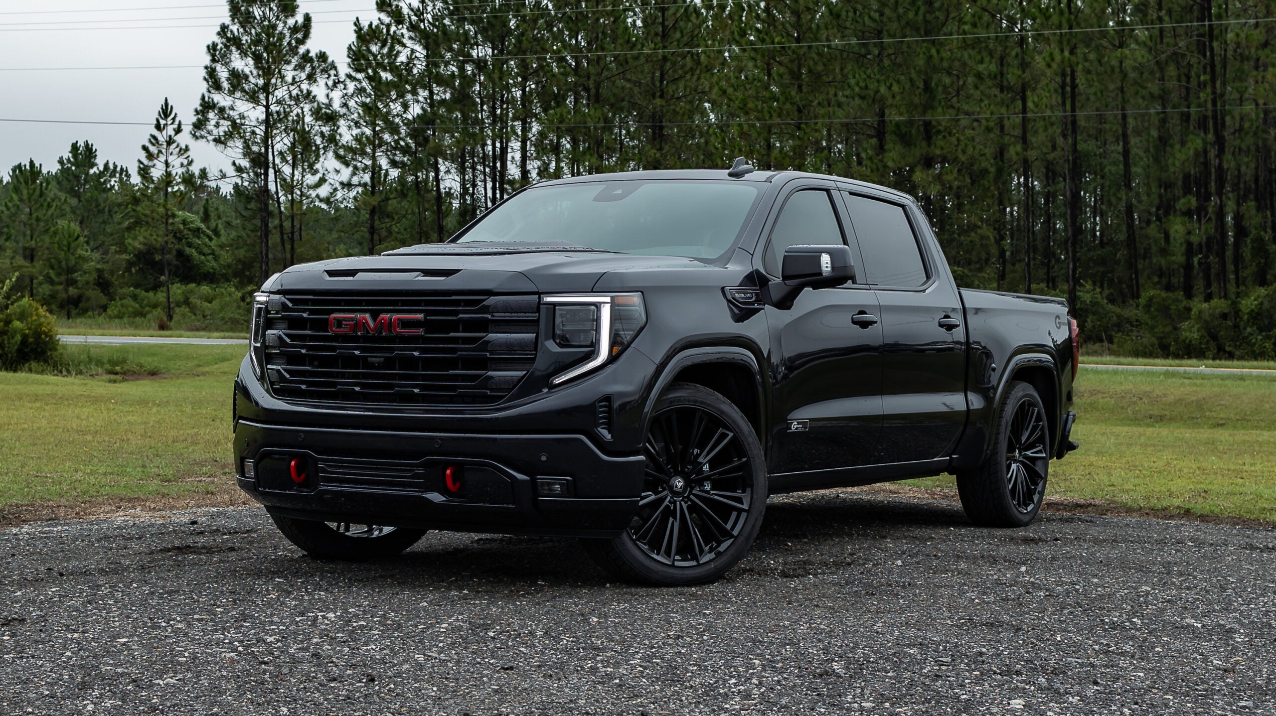 Black GMC Sierra pickup truck with custom black wheels and red tow hooks, parked on a gravel surface with trees behind it on a cloudy day.