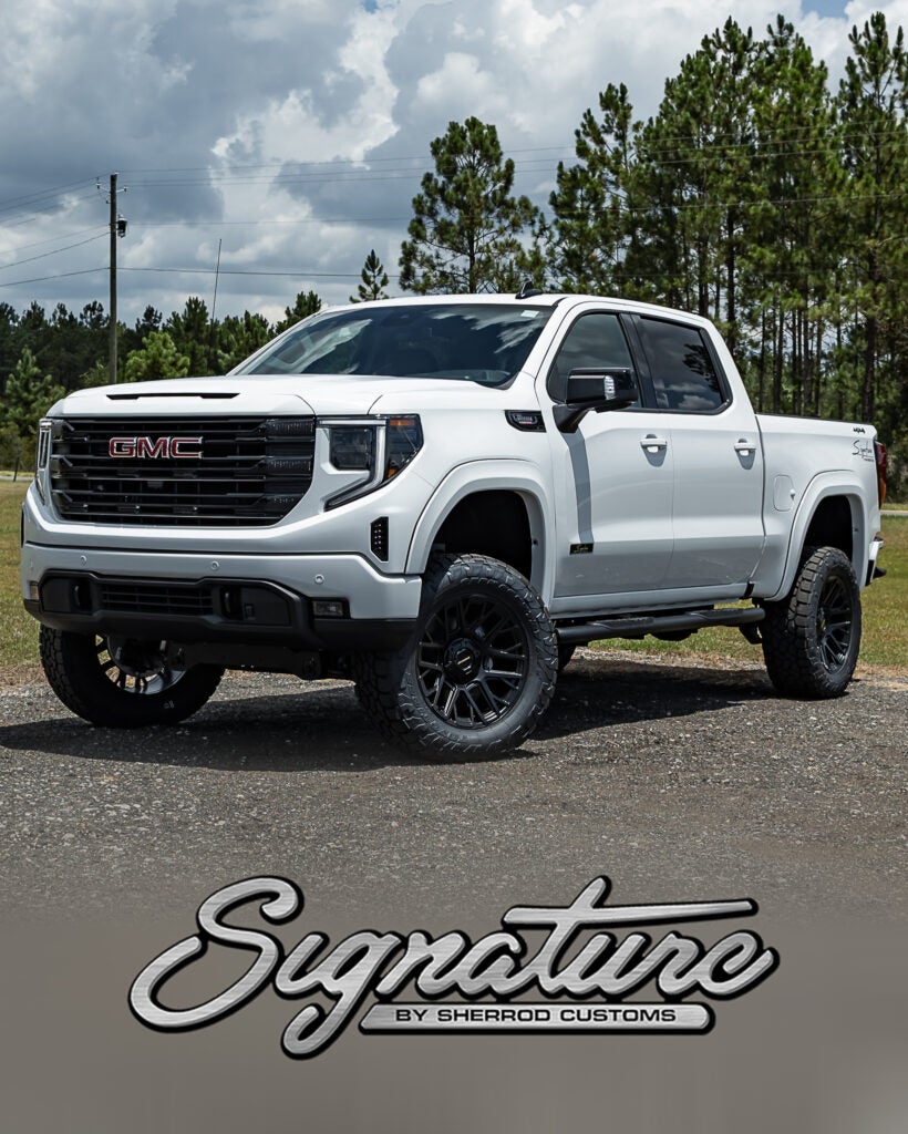 White lifted GMC Sierra pickup truck with black off-road wheels and side steps, parked outdoors with trees in the background and “Signature by Sherrod Customs” logo at the bottom.