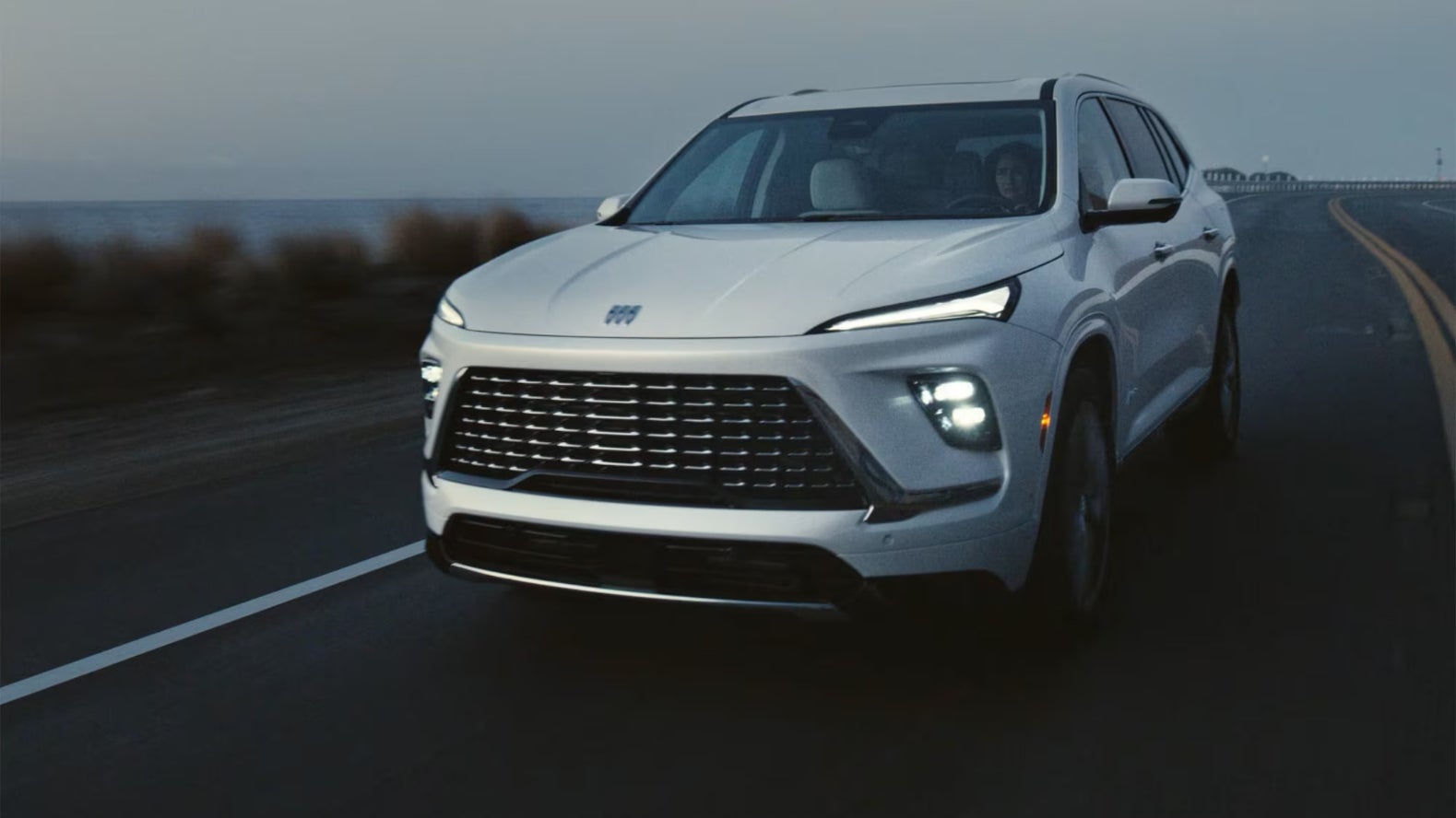 A front view of a white Buick Enclave driving down the road by a beach in cloudy weather.